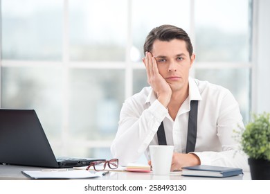 Young Businessman Is Bored While Working With Computer. Office Interior With Big Window. Man Looking At Camera