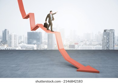 Young businessman with briefcase balancing on abstract falling red arrow on light city background with mock up place. Crisis, economy and work concept