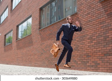 Young businessman with a briefcase and glasses running in a city street on a background of red brick wall