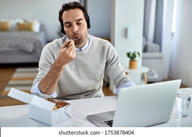 Young Businessman With Eyes Closed Enjoying In A Taste Of Food While Working On Laptop At Home. 