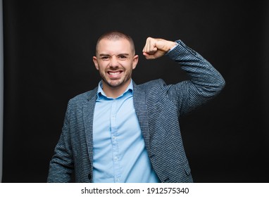 young businessman feeling happy, satisfied and powerful, flexing fit and muscular biceps, looking strong after the gym against flat wall