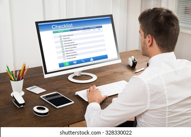 Young Businessman Filling Checklist Form On Computer At Wooden Desk