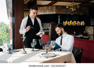 A young businessman in a fine restaurant examines the menu and makes an order to a young waiter in a stylish apron. Customer service. Table service in the restaurant. - Powered by Shutterstock