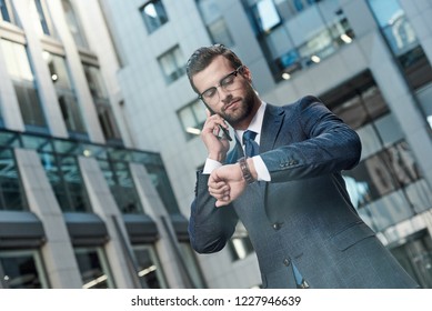 A young businessman with glasses and a beard negotiates a meeting. He has a smartphone in his hands and looks at his watch. - Shutterstock ID 1227946639