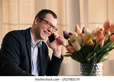 Young businessman in glasses and suit is talking with his beloved woman on mobile phone against the backdrop of bouquet of tulips. He arranged surprise for her. March 8th, birthday or Valentine's Day.