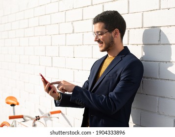A young businessman goes to work by bike using his mobile cell phone leaning against the white brick wall of the city - Shutterstock ID 1904333896