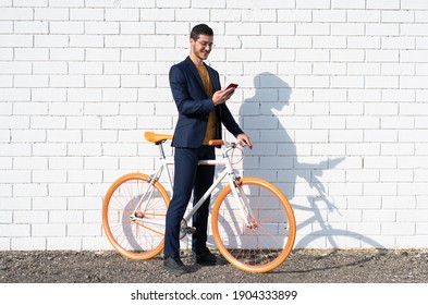 A Young Businessman Goes To Work By Bike Using His Mobile Cell Phone Leaning Against The White Brick Wall Of The City