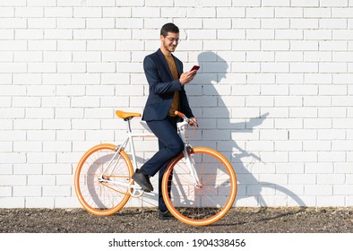A young businessman goes to work by bike using his mobile cell phone leaning against the brick wall of the city.