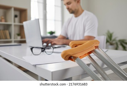 Young Businessman Has Injury And Uses Crutches. Close Up Of Crutches On Background Of Concentrated Man Working On Laptop In Office. Medical Equipment And Business Concept. Selective Focus.