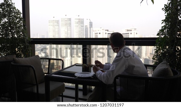 A young\
businessman is having breakfast in the terrace of the restaurant\
overlooking the skyscrapers.