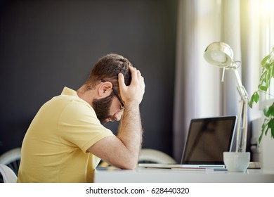 Young Businessman Having A Headache In Home Office While Sitting At The Table. Hands On Head, Computer In Front Of Him