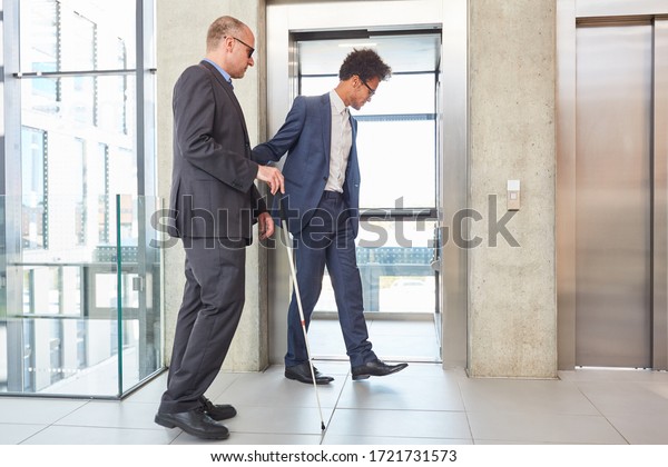 Young businessman helps a blind man with a cane\
at the elevator