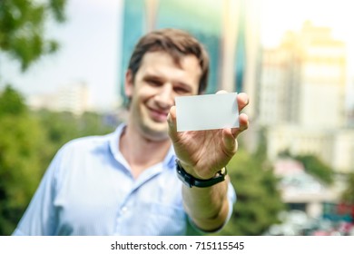 Young Businessman Holding Blank Visit Card In Hand And Smiling With Outdoor Trees And Office Building. Hand Is In Focus.