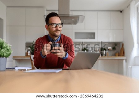 Young businessman holding coffee cup and contemplating while sitting with laptop on desk at home