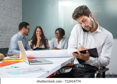 Young businessman holding mobile phone between ear and shoulder talking and writing notes to clipboard in modern office, while his business partners having meeting in the background.