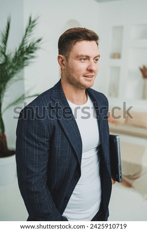 A young businessman in a jacket with a laptop stands in a bright room and works remotely. Work from home. A freelancer works online on a laptop.