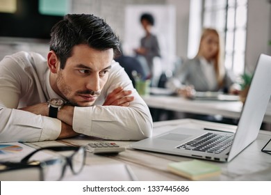 Young businessman leaning on his desk and feeling bored while surfing the net on a computer at work. There are people in the background.