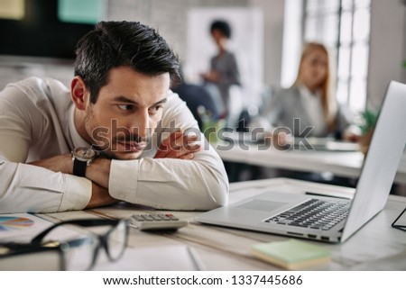 Young businessman leaning on his desk and feeling bored while surfing the net on a computer at work. There are people in the background.