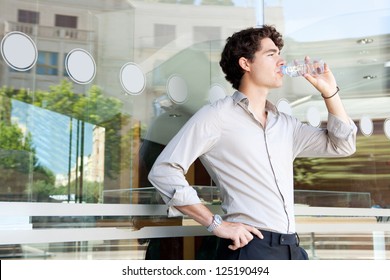 Young businessman leaning on an office building glass window drinking from a bottle of mineral water, with reflections of the city behind him.