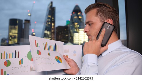 Young Businessman Looking On Pie Chart Sales Report While Talking On Mobile Phone In London Skyline City Center