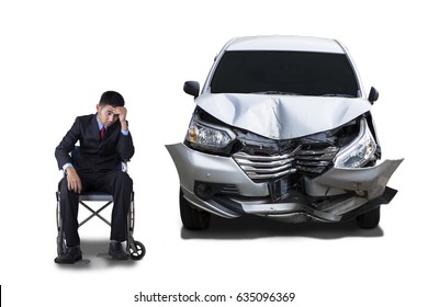 Young Businessman Looks Stressful, Sitting On Wheelchair Next To A Damaged Car. Isolated On White Background