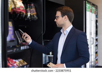 Young businessman makes quick cashless payment, lean mobile phone to card reader machine. Client buy goods in professional vending machine using modern technology. Retail place, mobile app, lifestyle