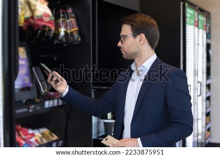 Young businessman makes quick cashless payment, lean mobile phone to card reader machine. Client buy goods in professional vending machine using modern technology. Retail place, mobile app, lifestyle