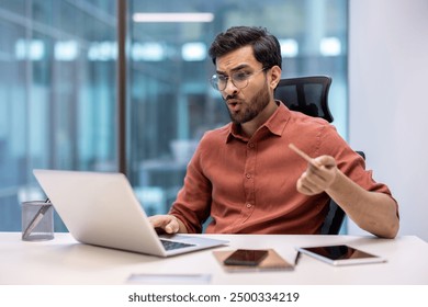 Young businessman in office reacts with surprise to content on laptop screen. Scene includes desk, tablets, notebook, phone, and office supplies. - Powered by Shutterstock