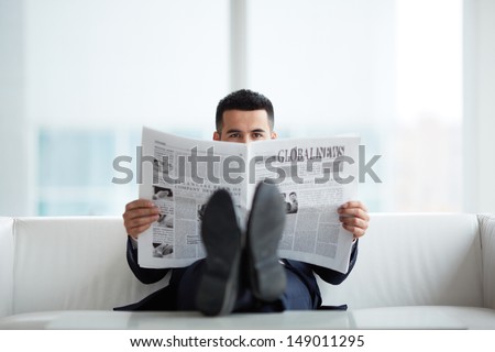 A young businessman on sofa looking at camera while reading newspaper