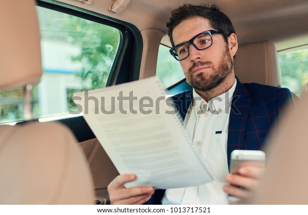 Young businessman reading paperwork and using a\
cellphone while sitting in the backseat of a car being driven\
through the city