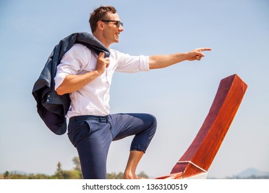 A Young Businessman In A Shirt With A Laptop Sits On A Boat And Points Into The Distance. Full Speed Ahead. Happy Businessman On Vacation.