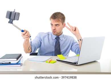 Young Businessman In Shirt And Tie Sitting At Office Computer Desk Holding Selfie Stick Shooting Self Portrait Photo Or Recording Video With Mobile Phone Acting Clown And Funny