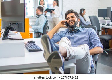 Young Businessman Sitting In An Office With His Legs Up On The Desk, Talking At His Cellphone.