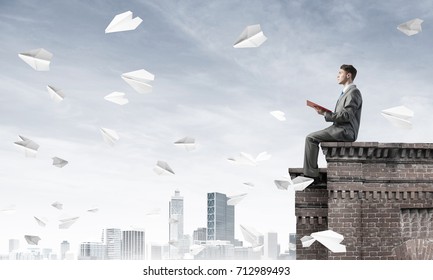 Young Businessman Sitting On Building Top With Red Book In Hands