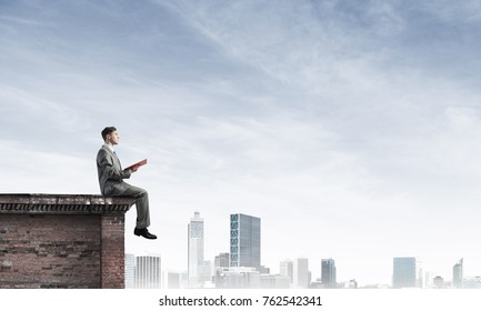 Young Businessman Sitting On Building Top With Red Book In Hands