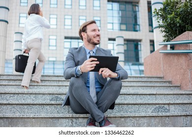 Young businessman sitting on stairs in front of an office building, working on a tablet. Woman climbs the stairs, talking on the phone, blurred in background. Work anywhere concept. - Powered by Shutterstock