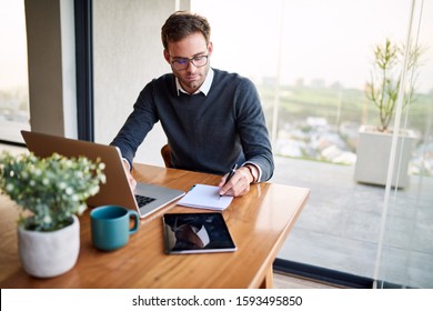 Young businessman sitting at a table at home working on a laptop and writing notes down on a notepad