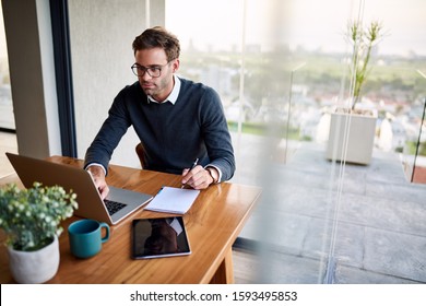 Young businessman sitting at a table at home working on a laptop and writing down ideas in a notebook - Shutterstock ID 1593495853