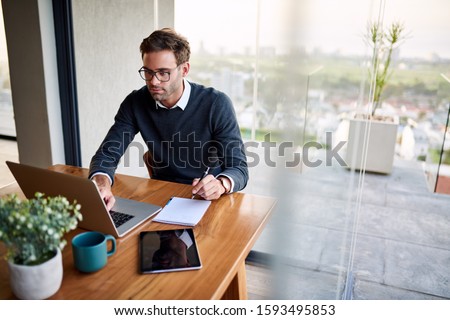 Young businessman sitting at a table at home working on a laptop and writing down ideas in a notebook