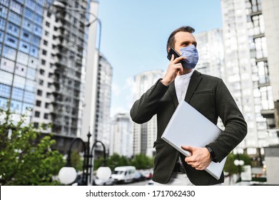 A young businessman in smart casual is walking in cases during pandemic. A guy is in medical mask with laptop talking on the phone outdoors among skyscrapers - Powered by Shutterstock