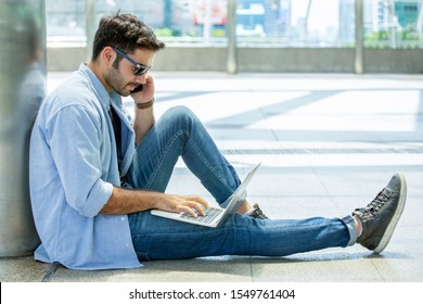 young businessman in smart casual wear sitting on street using laptop computer and talking by mobile phone in urban city outdoors. freelancer working on floor outside calling to customer. blue jeans