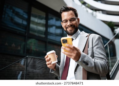 Young businessman is smiling while using smartphone and holding coffee outside modern office building