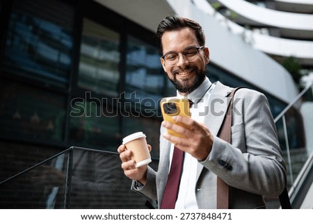 Young businessman is smiling while using smartphone and holding coffee outside modern office building