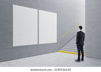 Young Businessman Standing In Creative Light Concrete Tile Interior With Stairs And Mock Up Poster On Wall. School Hallway And Corridor Concept