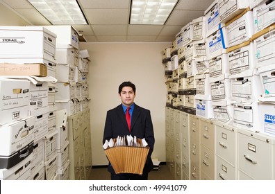 A young businessman is standing in a file room holding files. Cabinets and storage boxes line the sides. Horizontal shot. - Powered by Shutterstock