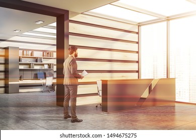 Young businessman standing in modern office with gray and wooden walls, grey reception desk and workplace with computer in background. Toned image