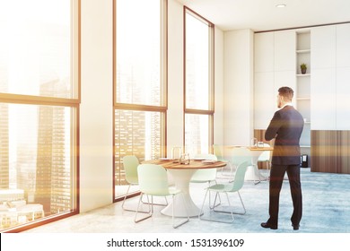 Young Businessman Standing In Modern Office Canteen With White Walls, Round Dining Tables With Blue Chairs And Bookcase. Toned Image