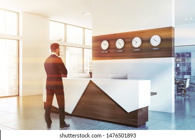 Young businessman standing near white and wooden office reception desk with computers and clocks showing world time. International company concept. Toned image