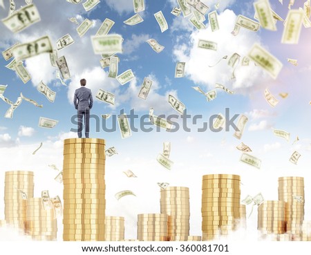 Young businessman standing on a pile of golden coins, proud of himself, other piles around, dollars falling from above. Blue sky at the background. Concept of earning money and prosperity.