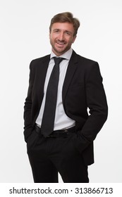 Young Businessman Standing In Photo Studio With His Hands In Pockets. Blond Man In Business Suit Smiling For The Camera.